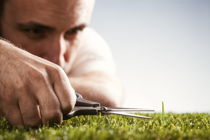 Photo of a grass lawn with one blade of grass longer than the rest. A man is on the ground with a pair of scissors to cut the single blade of grass to match the rest.