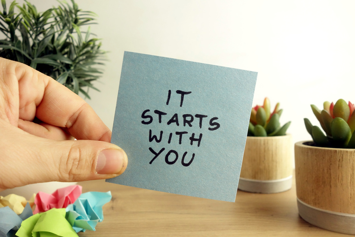 Photo of a hand holding a sticky note that says "it starts with you"