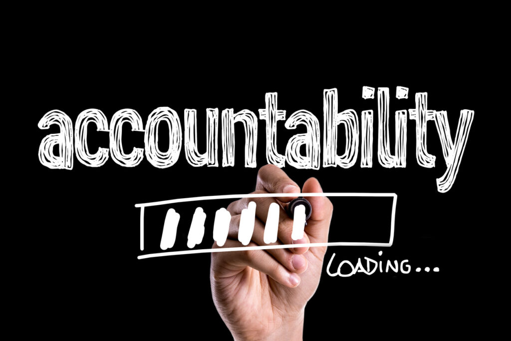 The image shows a black background with the word “accountability” written in large, white, chalk-style lettering across the top. In the foreground, a hand is holding a marker and appears to be drawing a loading bar underneath the word. The loading bar is partially filled with several white segments, and the text “LOADING…” is written to the right of it.