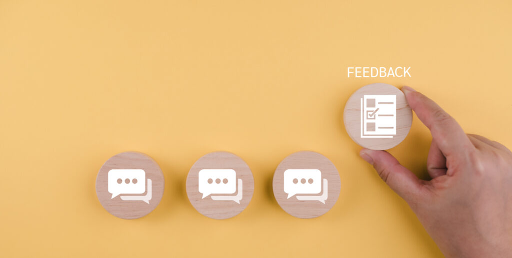 The image shows a bright yellow background with four small, round wooden tokens arranged in a row. Three tokens on the left display white speech-bubble icons, suggesting conversation or messages. On the right, a hand is placing or holding a fourth wooden token that shows a checklist icon, with the word “FEEDBACK” written above it. The overall visual suggests the idea of collecting comments or turning conversations into structured feedback.