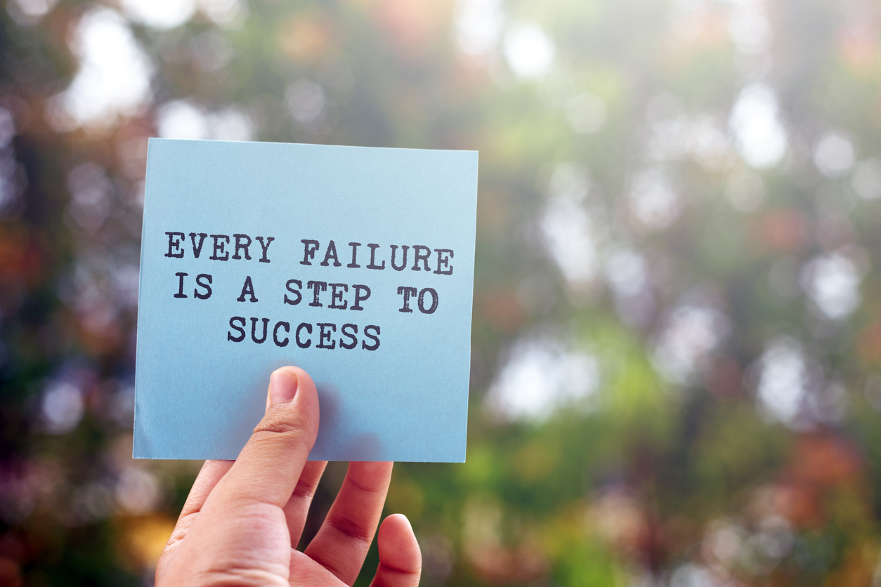A hand holding up a sticky note that says "every failure is a step to success" against a blurred colorful background