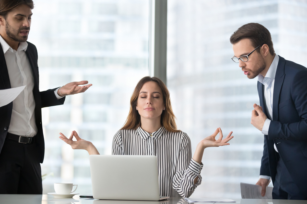 A woman is seated at a desk in front of an open laptop. She has her eyes closed and her hands raised slightly to either side, palms up, as if pausing, centering herself, or tuning out distractions. Two men in business attire stand on either side of her. Both appear to be talking or gesturing toward her—one holding papers and gesturing with his hand, the other leaning in slightly while also holding documents.