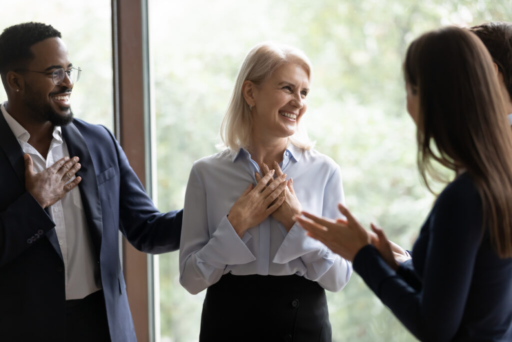 The image shows a small group of adults in a professional setting, standing indoors near a large window with greenery visible outside. They appear to be engaged in a warm, positive conversation. At the center is a woman with light-colored hair, smiling and holding her hands over her chest, suggesting gratitude or appreciation. She’s dressed in business attire. To her left, a man in a suit and glasses is smiling and also touching his chest, mirroring the appreciative gesture. On the right, another person (seen mostly from behind) is clapping or gesturing enthusiastically toward the woman in the center.