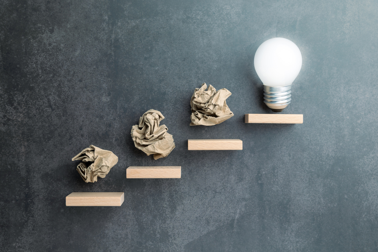 Conceptual image of crumpled paper balls as steps leading to a light bulb, symbolizing failure as part of the creative or problem-solving journey toward success