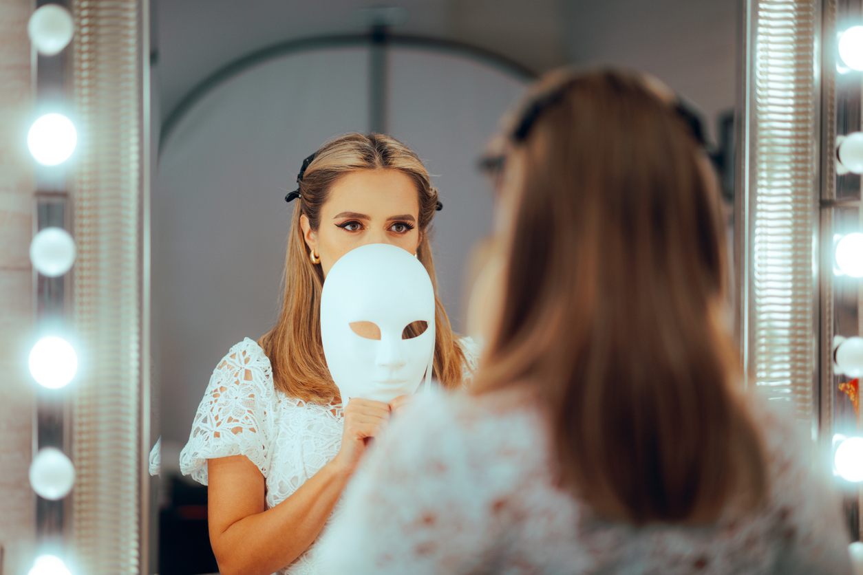A woman standing in front of a large, well-lit mirror, likely in a dressing room or backstage area. The mirror is framed with bright round vanity lights. She is holding a plain white mask up to her face, covering her nose and mouth, while looking at her reflection.
