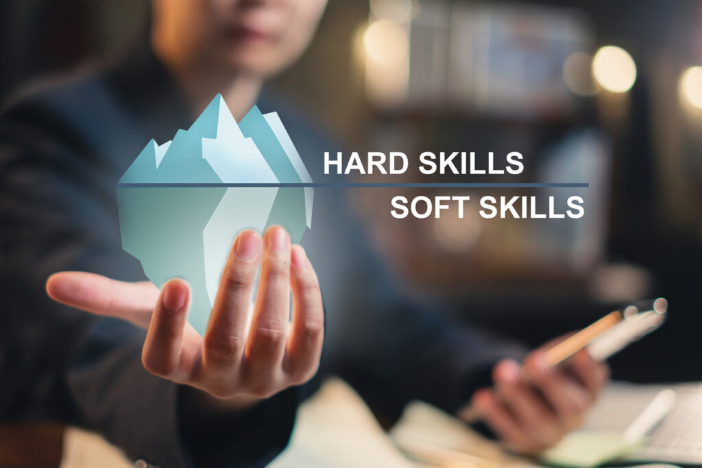 A person holding a stylized iceberg graphic labeled “hard skills” above the surface and “soft skills” below, illustrating that soft skills are deeper and less visible.
