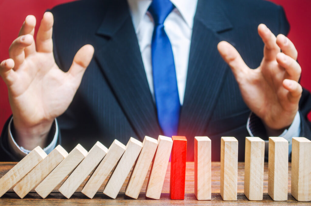 A person in a suit holds their hands over a row of domino blocks. Some dominoes on the left are falling, but a red domino in the middle stops the chain reaction, leaving the dominoes on the right standing.