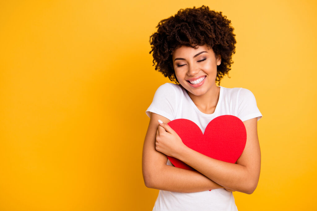 A smiling woman with curly hair hugs a large red heart against a bright yellow background, eyes closed in a joyful, affectionate pose.
