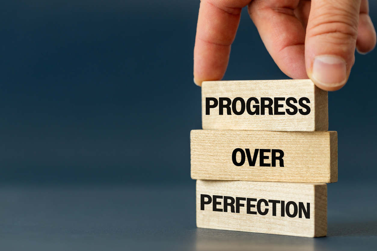 A hand places a wooden block on top of a stack that reads “PROGRESS OVER PERFECTION,” emphasizing steady improvement rather than flawlessness.