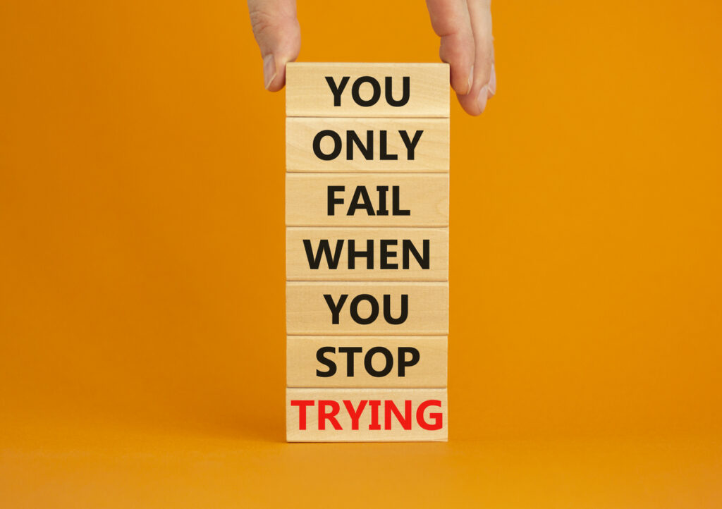 A hand stacking wooden blocks that say "you only fail when you stop trying", set against an orange background