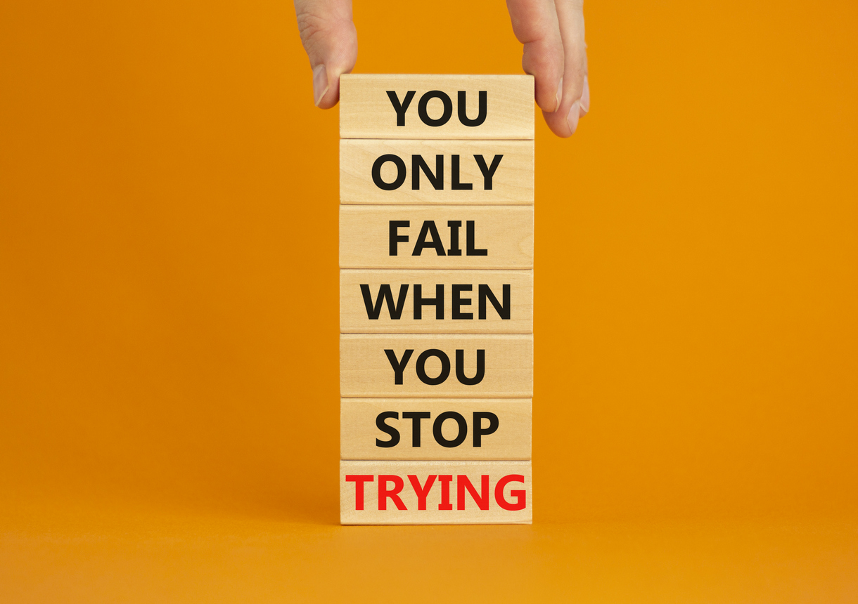 A hand stacking wooden blocks that say "you only fail when you stop trying", set against an orange background