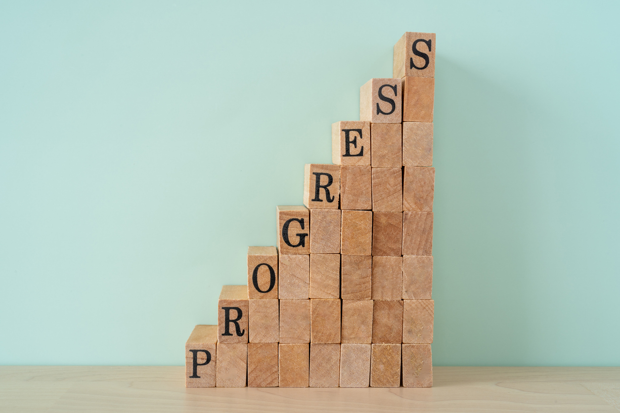A staircase of stacked wooden blocks rises from left to right, with letters on select blocks spelling “PROGRESS” diagonally upward against a simple light green background. The image symbolizes gradual improvement, growth, and step-by-step achievement.
