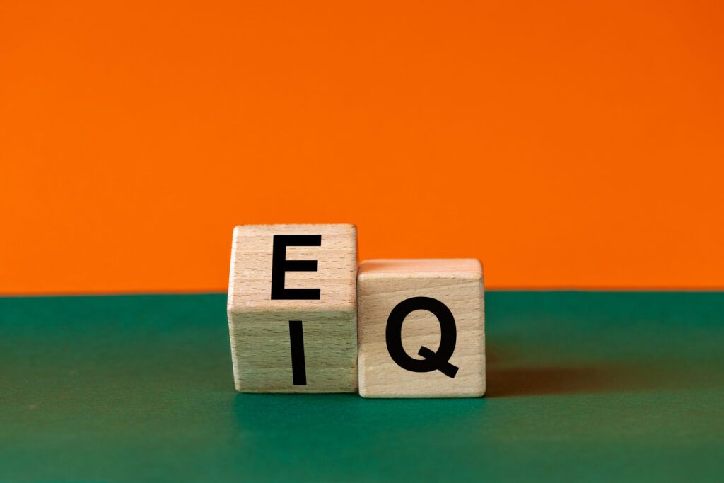 Two wooden letter blocks arranged to spell “IQ,” sitting on a green surface against a bright orange background. The “I” and “Q” are visible on the front faces, with an “E” visible on the top/front of the left block.