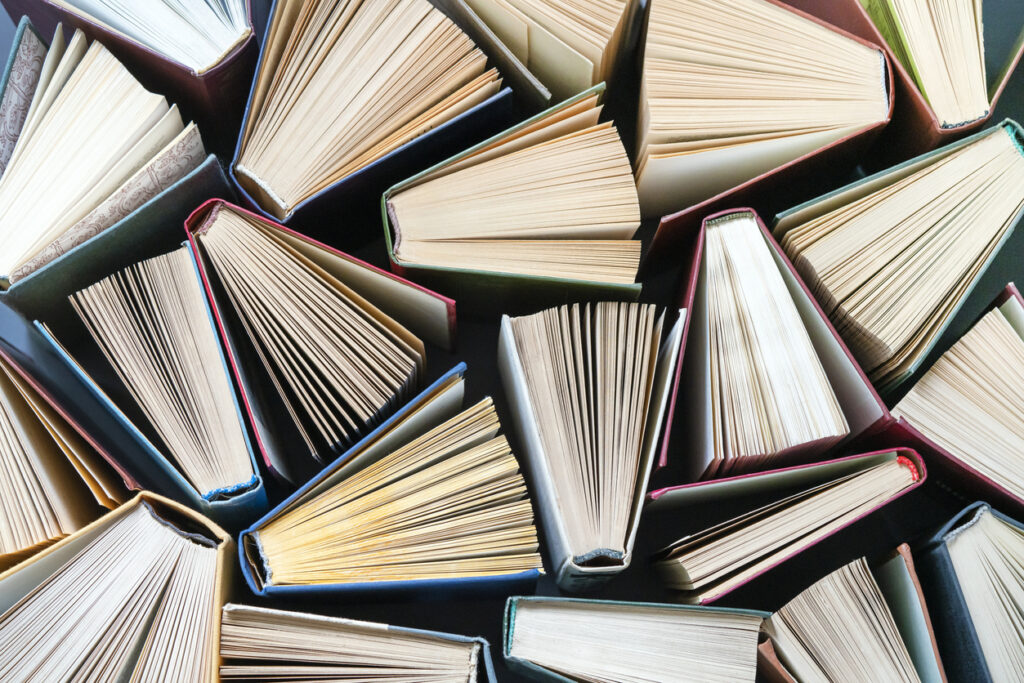 A top-down view of numerous hardcover books arranged closely together, with their pages fanned open in different directions, creating a textured, overlapping pattern of cream-colored pages and colorful book covers.