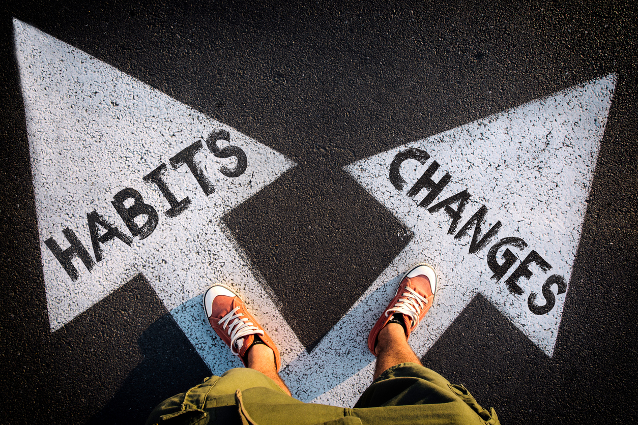 A first-person view of someone standing at the junction of two large painted arrows on pavement, one labeled "HABITS" pointing left and the other "CHANGES" pointing right, symbolizing a choice between staying the same and embracing change.