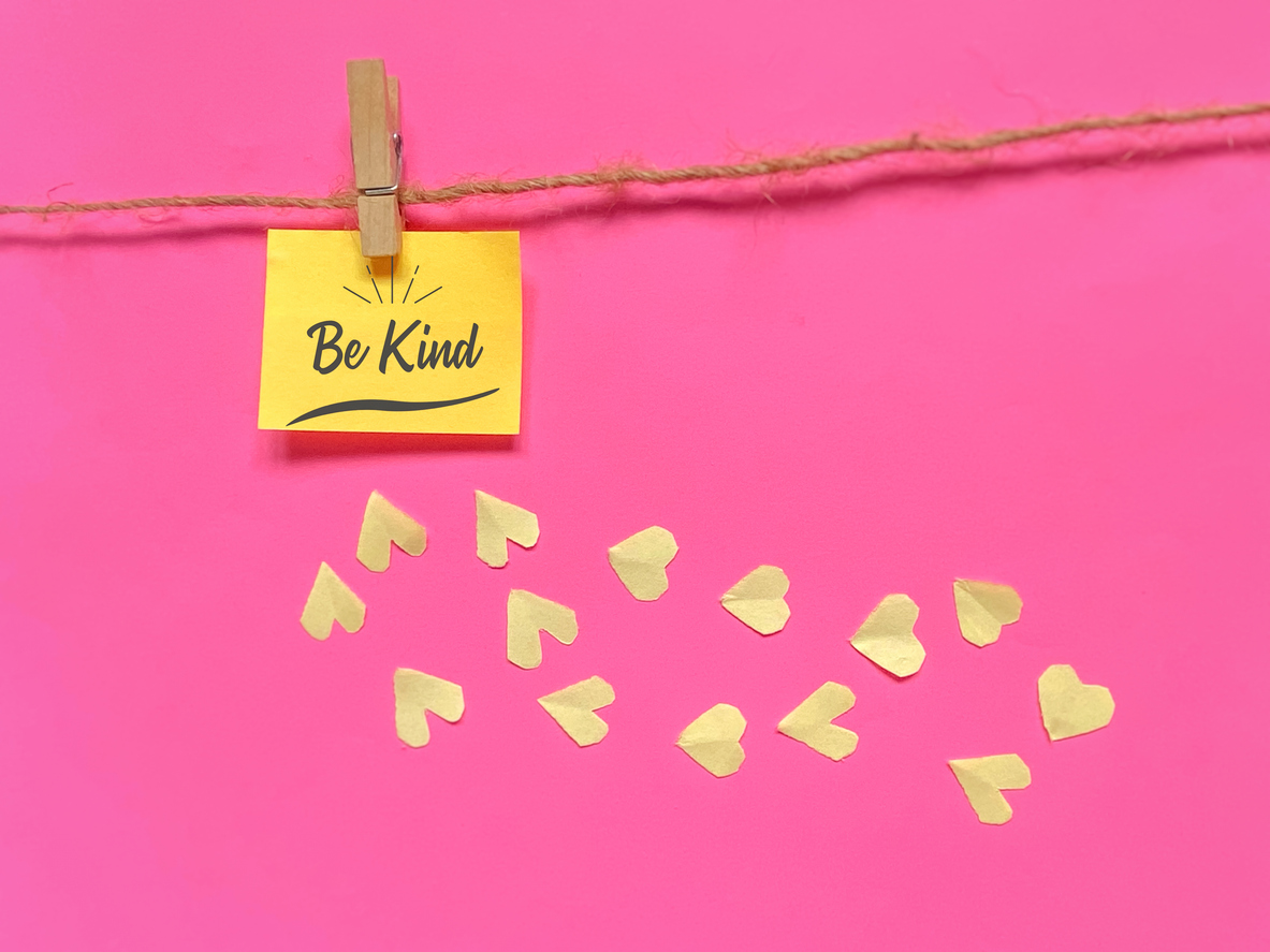 A yellow sticky note reading “Be Kind” is clipped to a string against a bright pink background, with small paper heart cutouts scattered below, symbolizing kindness, compassion, and positivity.