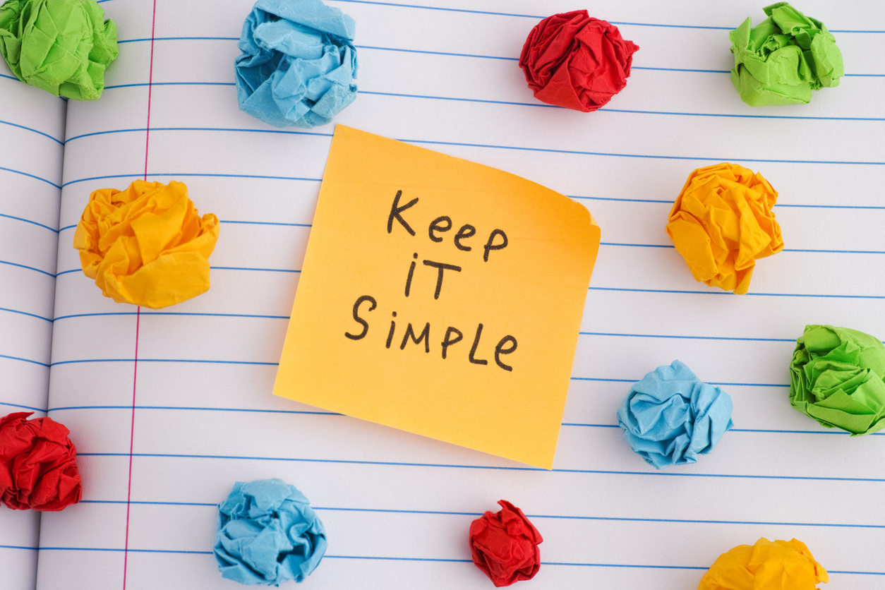 Keep it simple. A yellow paper note with the phrase Keep it simple on it with some colorful crumpled paper balls around it.