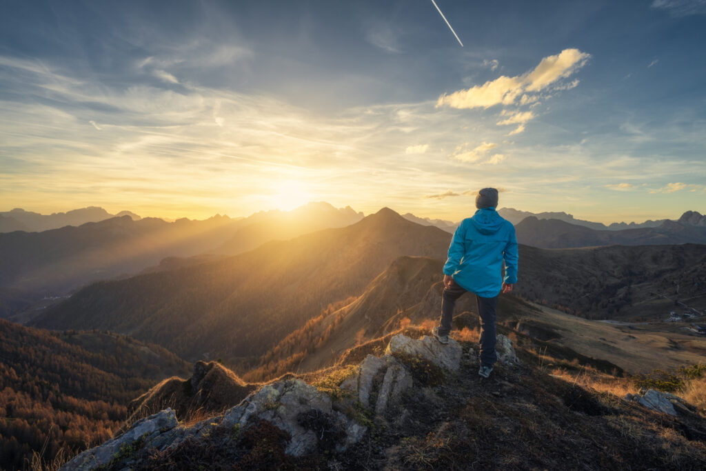 A hiker in a blue jacket stands on a rocky mountain ridge overlooking a dramatic range at sunset.