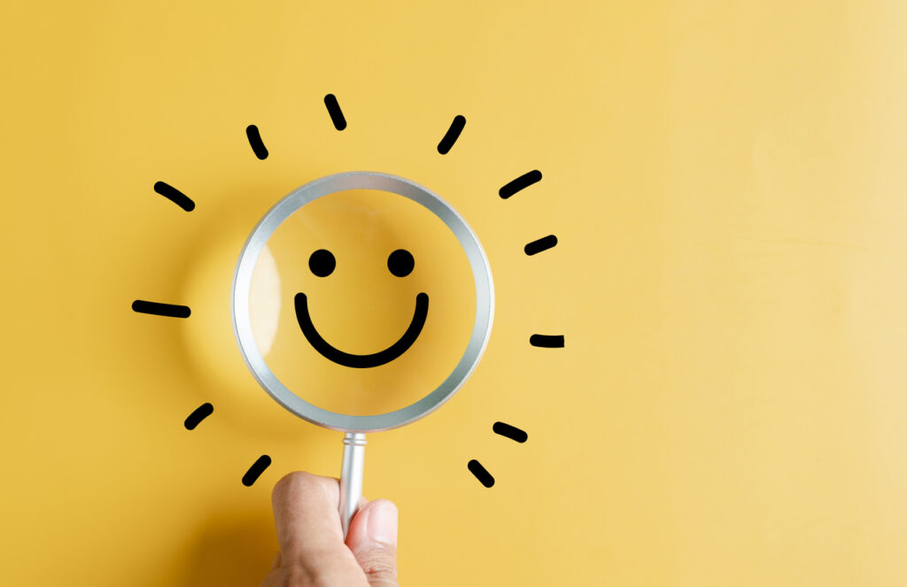 A magnifying glass with a smiling face is held over a bright yellow background, symbolizing positivity and finding happiness.