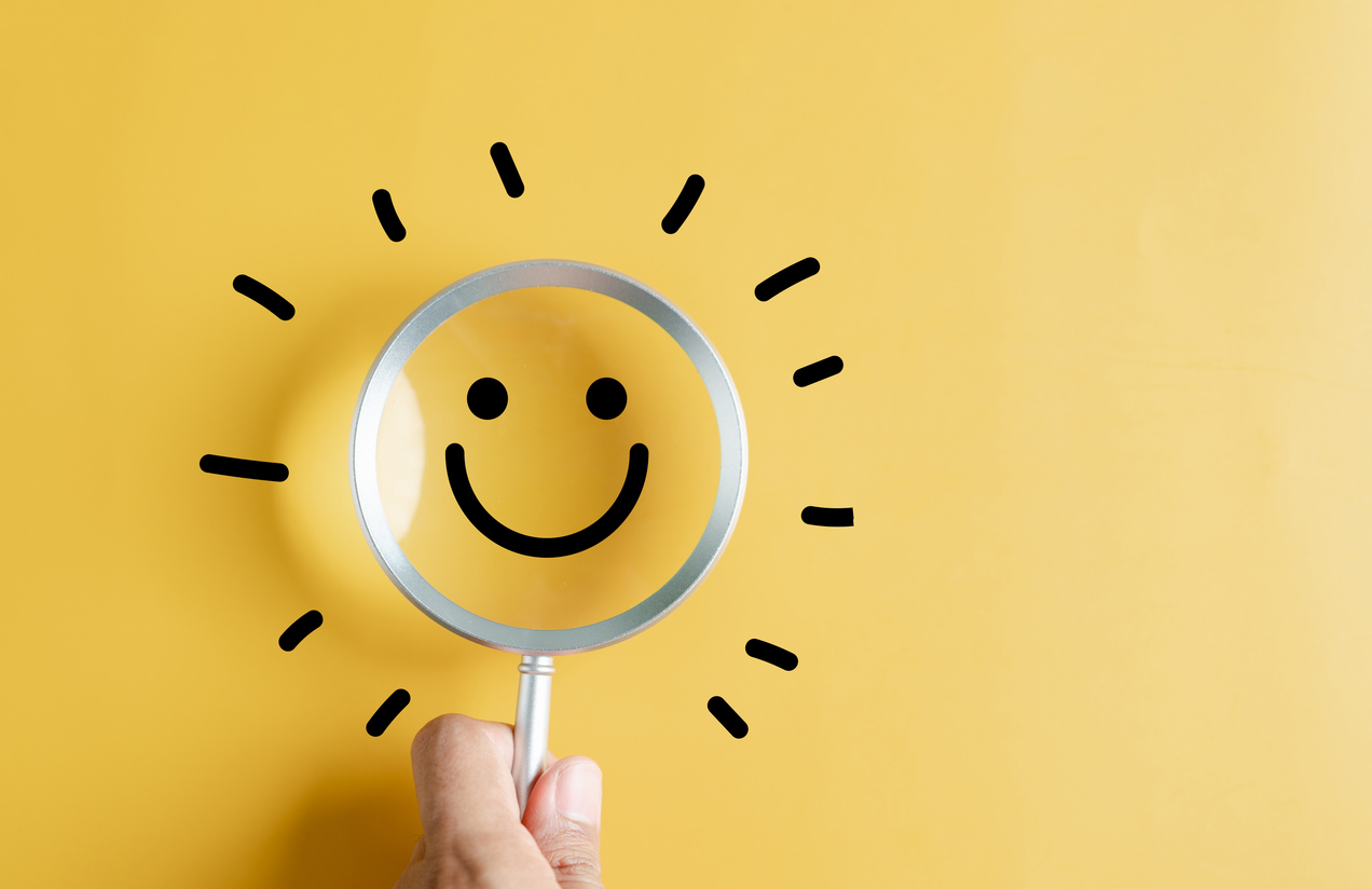 A magnifying glass with a smiling face is held over a bright yellow background, symbolizing positivity and finding happiness.