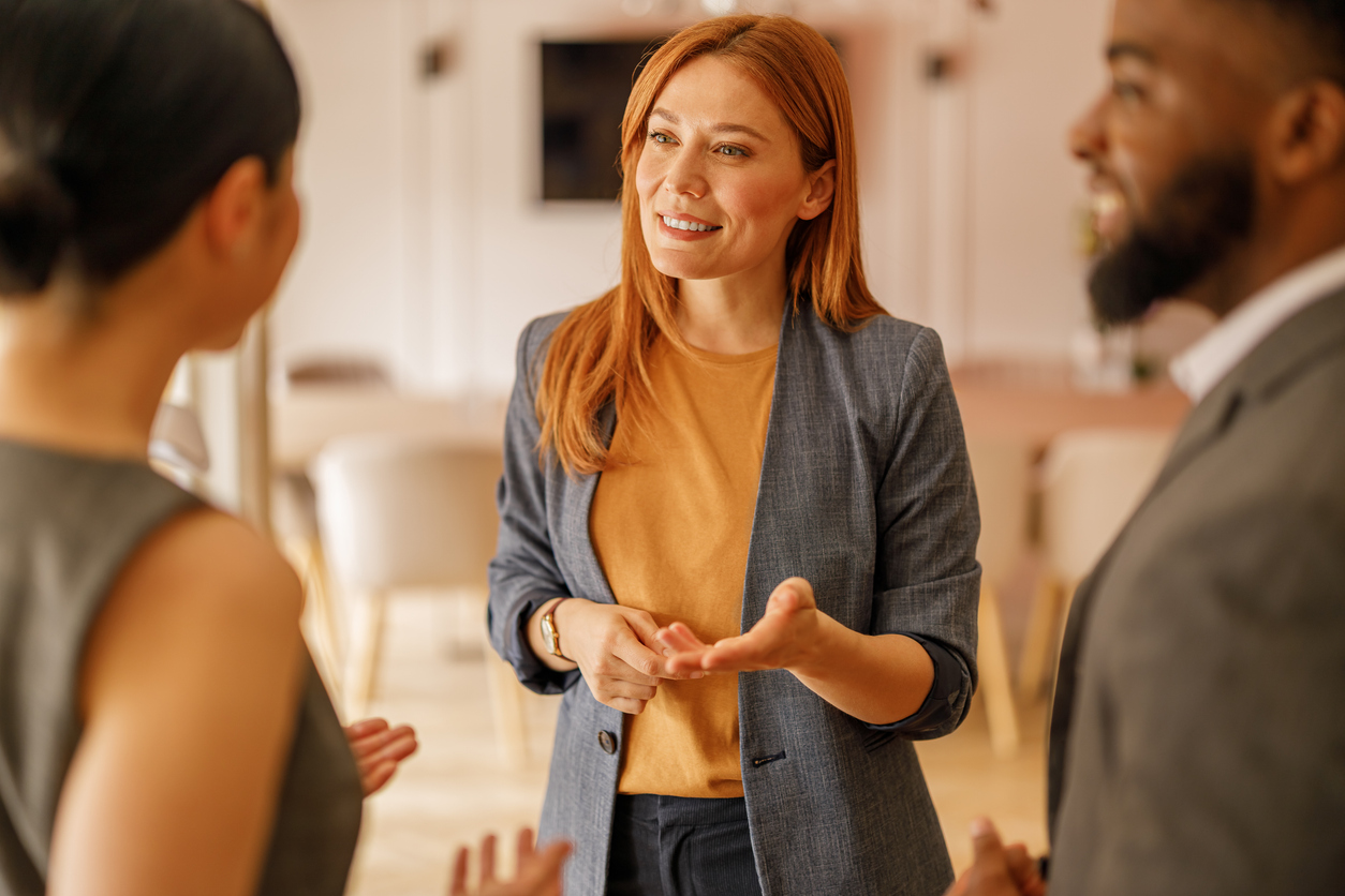 A red-haired woman in a gray blazer gestures while speaking with two colleagues in a warmly lit office, suggesting a friendly and engaging professional conversation.