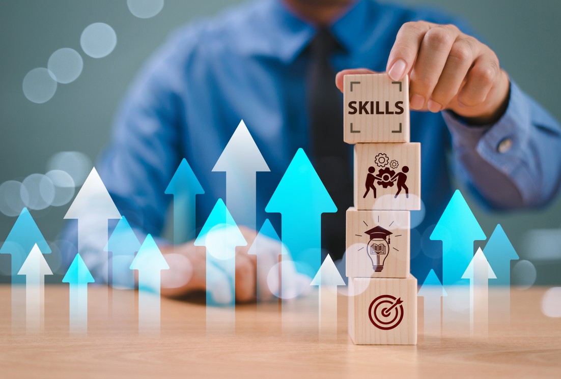 A business professional stacks wooden blocks labeled with skills, teamwork, education, and goals, while blue and white upward arrows symbolize career growth, continuous learning, and professional success.